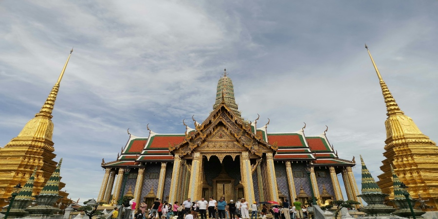Golden spires of Wat Phra Kaew gleam under blue skies, surrounded by lush greenery, epitomizing Thailand's rich cultural heritage and spiritual significance.